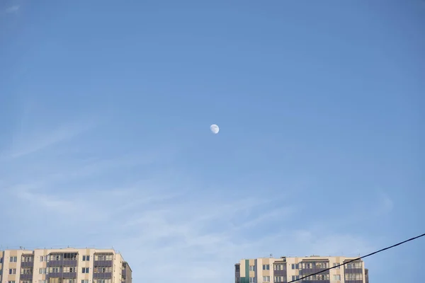 Moon on bright blue sky with light feathery clouds and tops of h