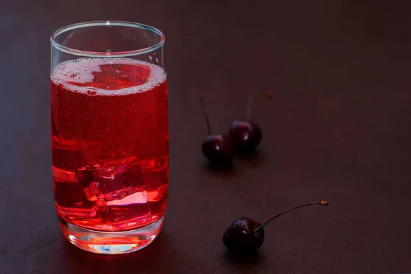 Beba com cerejas geladas, cerejas doces em um fundo escuro. Cocktail de ameixa fresco. Cocktail fresco de verão com cerejas e cubos de gelo. Um copo de refrigerante beber uma cereja em um fundo escuro . — Fotografia de Stock