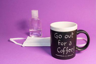 Tebeşirle yazılmış bir fincan kahve bir şişe dezenfekte jeli ve mor arka plandaki hastalıklara karşı koruyucu bir maskenin yanında. Yeni normallik, koruma veya enfeksiyon korkusu kavramları