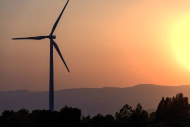 Dağları olan yel değirmeni kulesi ve gün batımının turuncu gökyüzü ya da arka planda gün doğumu ve ön planda ağaçlar. Kopyalama alanı olan. Temiz enerji, iklim değişikliği ve ekoloji kavramları.
