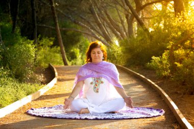 Nilüfer pozisyonunda oturmuş meditasyon yapan bir kadın ellerini Gyan Mudra 'da dizlerinin üstüne çöküp günbatımında ormanda bir patikada oturuyor.