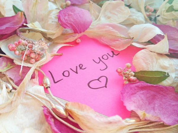 爱你的消息粉红色的便条与干燥的玫瑰和兰花花瓣和珠宝戒指和链在木桌背景 — 图库照片