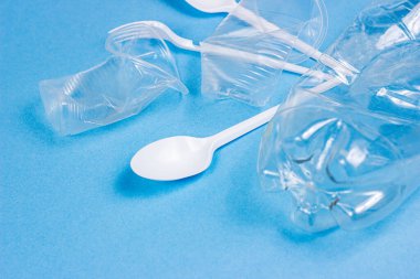 Ezilmiş plastik kaşıklar, çatallar, şişeler ve bardaklar parlak mavi arka planda tek kullanımlık atıklar. Çevre kirliliği ve çöp geri dönüşümü kavramı.