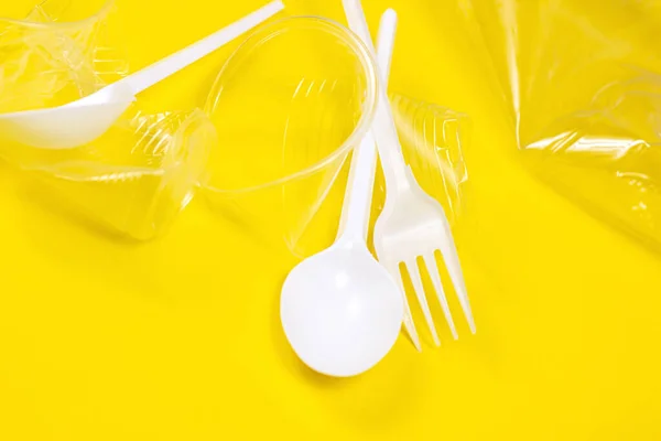 Crushed plastic spoons, forks, bottles and cups as a disposable waste on bright yellow background. Environmental pollution and litter recycling concept.