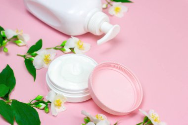Nemlendirici krem için beyaz plastik kozmetik konteynır. Fotokopi alanı olan açık pembe arka planda parlak yasemin çiçekleri. Vücut ve cilt bakımı, sağlık, sağlık ve güzellik konsepti.