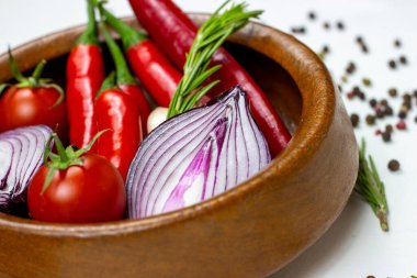 Parlak yaz sebzeleri: soğan, sarımsak, domates, kırmızı biber, kahverengi ahşap kasede biberiye ve beyaz masa arkasında biber..