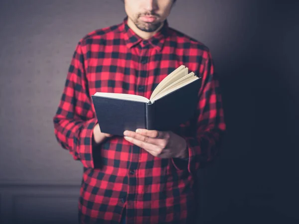 A young man wearing a red flannel shirt is reading a book in a luxury living room