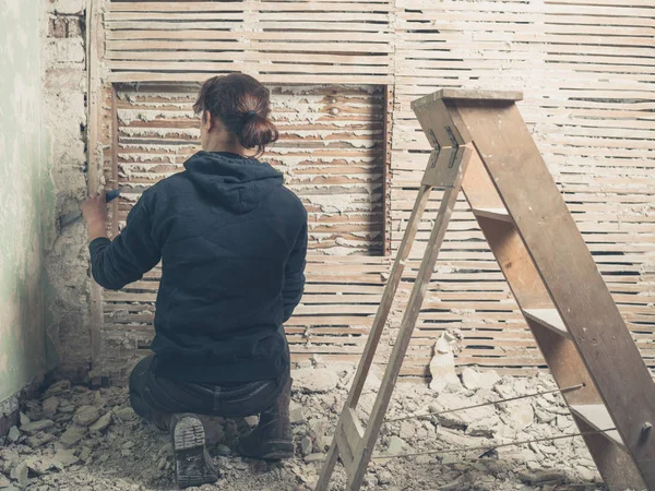 一个年轻的女人正在用锤子把石膏从和涂抹的墙上砍下来 — 图库照片