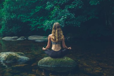 Genç bir kadın nehirdeki bir kaya üzerinde meditasyon