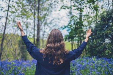 Genç bir kadın bluebells bir çayırda oturuyor ve sevinç kollarını kaldırarak dile getiriyor