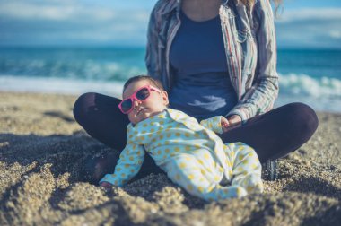 Serin bir bebek güneş gözlüğü takıyor ve annesi ile sahilde chilling