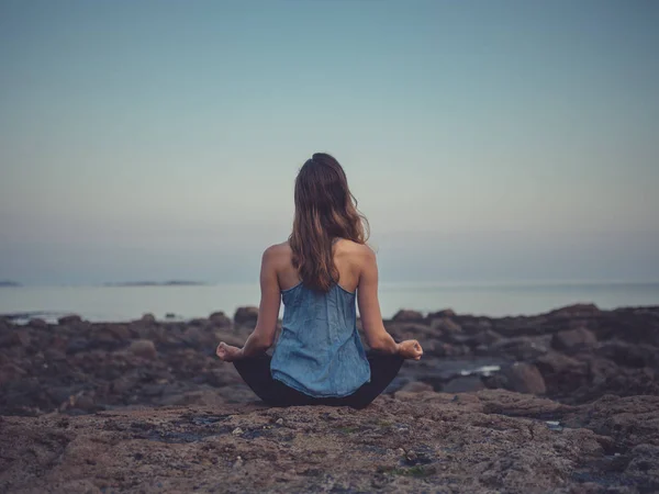 在日落的时候 年轻女子的后景坐在海边的岩石上冥想 — 图库照片