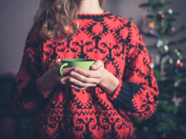 A young woman in a woollen jumper is drinking tea by the christmas tree