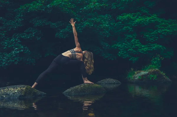 一个年轻的女人在河里的岩石上做瑜伽 她摆出了战士的姿势 — 图库照片