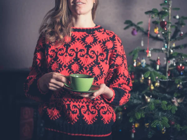 A young woman in a woollen jumper is drinking tea by the christmas tree