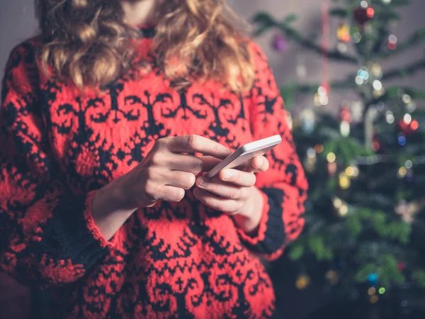 クリスマス ツリーで若い女性が彼女のスマート フォンを使用してください — ストック写真