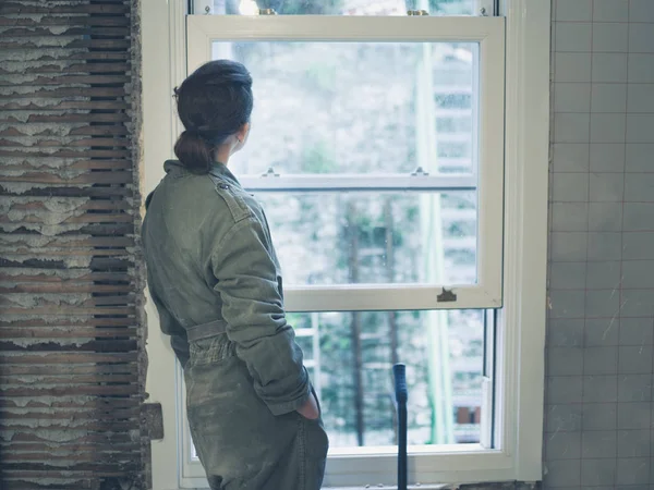 A young woman wearing a boiler suit is standing by the window and is day dreaming