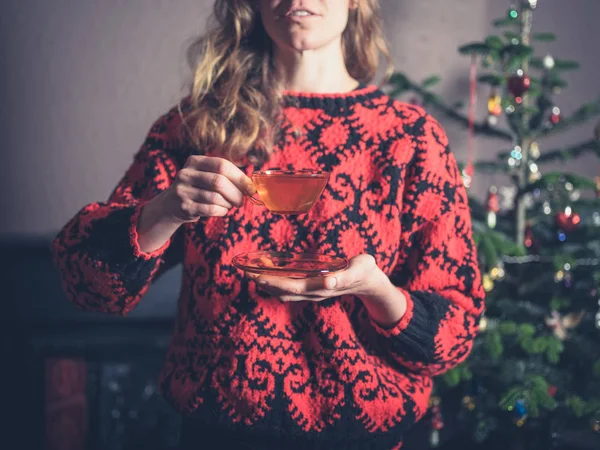 A young woman in a woollen jumper is drinking tea by the christmas tree