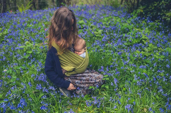 A young mother with a sleeping baby is sitting in a meadow of bluebells