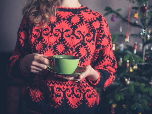 A young woman in a woollen jumper is drinking tea by the christmas tree