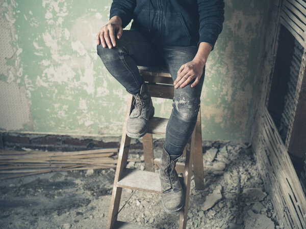 The legs of a young person sitting on a stepladder in a messy and derelict room