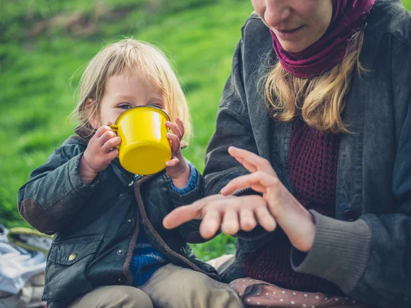 屋外で黄色いカップから飲む幼児の子供と近くに座っているお母さん — ストック写真