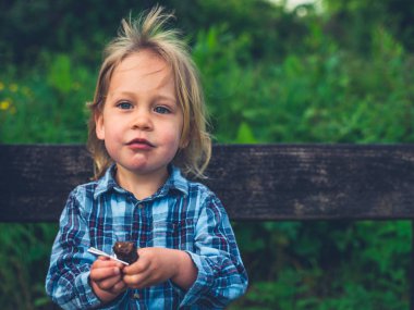 Küçük bir bebek doğada bir bankta oturuyor ve bir meyve bar yiyor