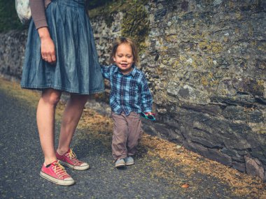 Genç bir anne ve yeni yürümeye başlayan çocuğu taş bir duvarın yanından geçiyorlar.
