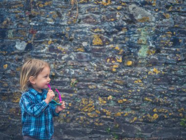 Güneş gözlüklerini tutan küçük bir çocuk taş bir duvarın yanında duruyor.