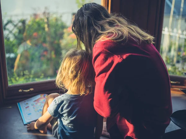 若い母親は日没時に窓際で幼児に本を読んでいる — ストック写真