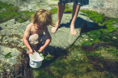 Küçük bir bebek annesiyle sahilde su ile bir kova dolduruyor
