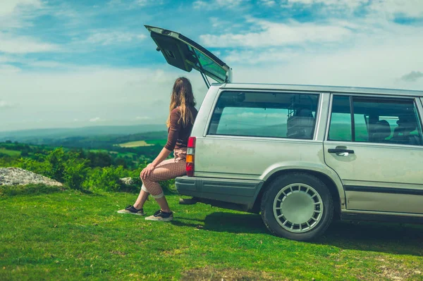 A young woman is sitting in the back of her stationwagon in nature on a sunny summer day