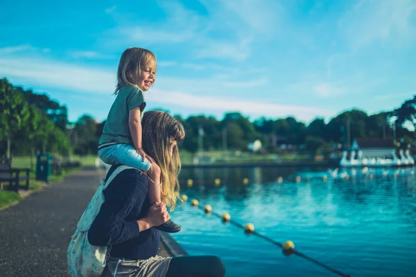 유아가 일몰에 공원에서 호수에 어머니의 어깨에 앉아있다 — 스톡 사진