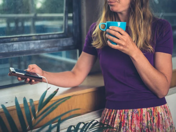 Ung Kvinna Dricker Kaffe Och Laddar Sin Telefon Genom Fönstret — Stockfoto