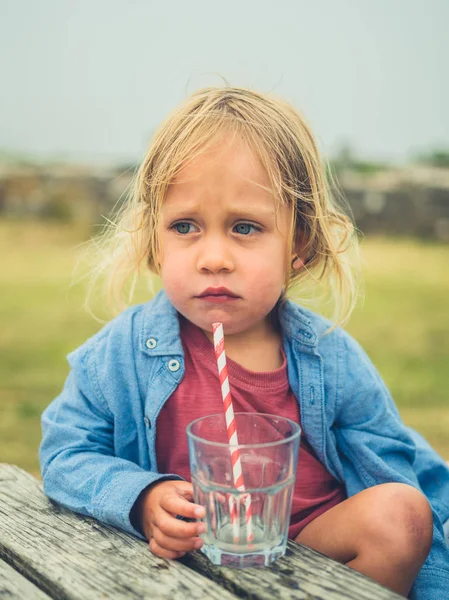 A little toddler is sitting at a picnic table outdoors and is drinking from a straw