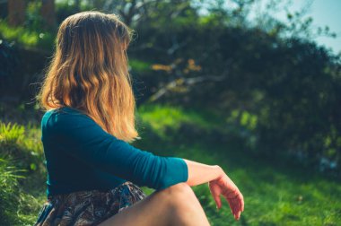 Genç bir kadın güneşli bir bahar gününde bahçesinde çimlerin üzerinde oturuyor.