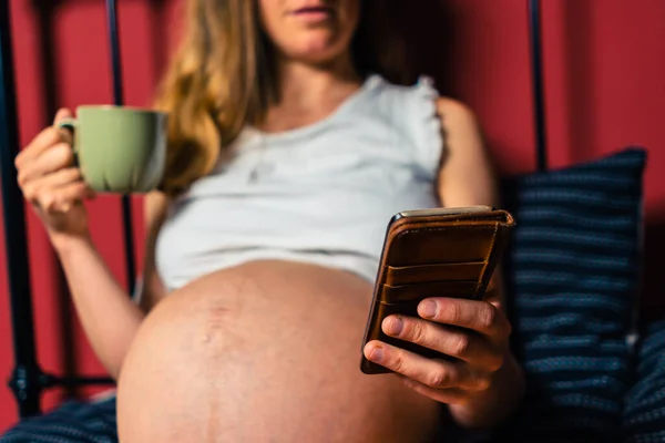 妊娠中の若い女性がマグカップから飲んでいて ベッドでスマートフォンを使っています — ストック写真