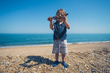Küçük bir anaokulu çocuğu elinde yosunla sahilde duruyor.