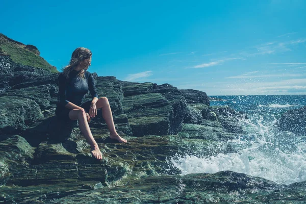 一位穿着湿衣的年轻女子正坐在海边的岩石上 这时海浪正冲进来 — 图库照片