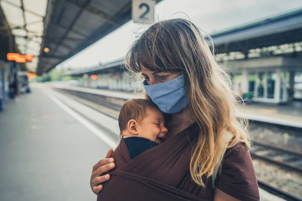 赤ん坊を抱えた若い母親が駅でフェイスマスクをしている — ストック写真