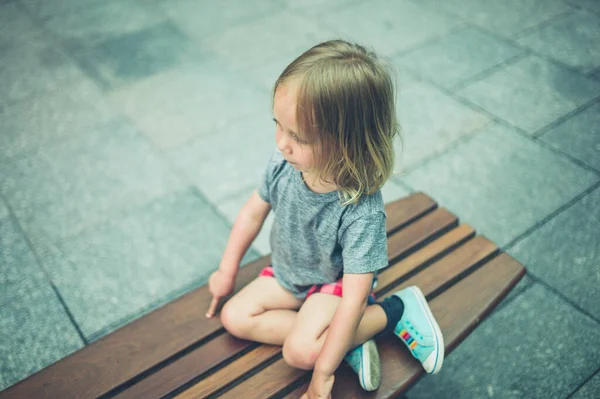 Küçük bir anaokulu öğrencisi şehirde bir bankta oturuyor.