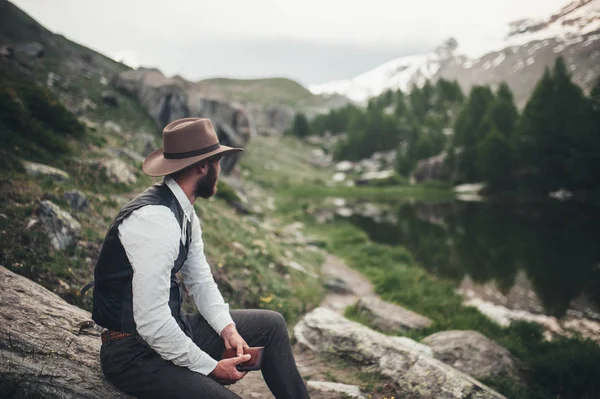 A man in a hat sits on the lake and looks at the landscape and holds a notebook in his hands
