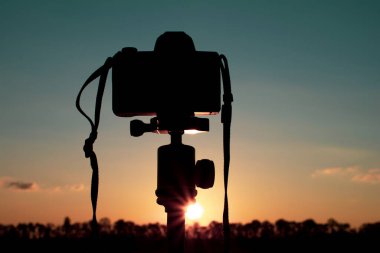 Çimlerde kırmızı bir günbatımı çeken üç ayaklı bir kameranın silüeti.