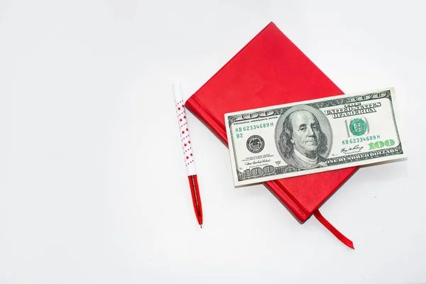 Beyaz arka planda 100 dolarlık kırmızı defter ve kalem.