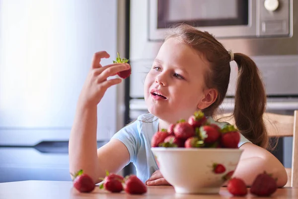 Hermoso niño europeo en la cocina comiendo fresas y bebiendo leche . — Foto de Stock