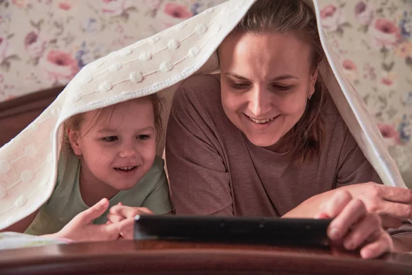 Une charmante petite fille et sa belle jeune mère utilisent une tablette numérique et sourient assis sur des chaises à la maison Image En Vente