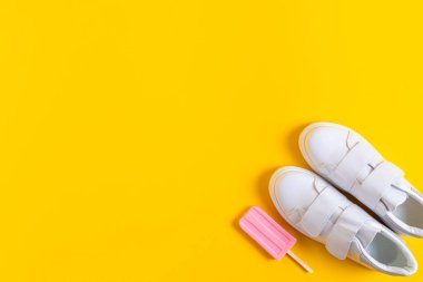 Çerçevenin köşesindeki sarı arka planda beyaz spor ayakkabılar ve pembe dondurma. Yukarıdan bak. Sıcak parlak yaz mevsimi konsepti