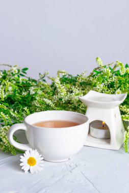 Taze kır çiçeklerinin arka planında beyaz bir fincan çay ve aromalamp. Dinlenme kavramı, rahatlama.