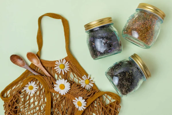 Flat lay of three glass jars with tea, wooden spoons and a reusable string bag on a mint background decorated with daisies. Reusable concept, environmental friendliness