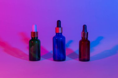 Neon arka planında damlatıcıları olan üç cam şişe. Arkaplan gradyanı pembeden maviye. İki gölge. Yatay fotoğraf. bakım kozmetik konsepti.